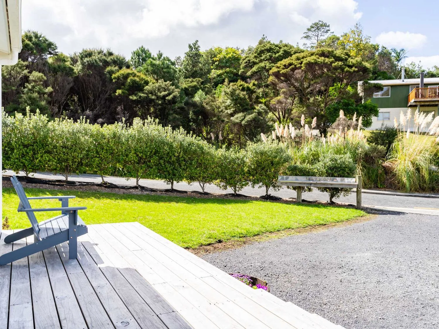 Una terraza de madera con una silla azul con vistas a un jardín con césped, arbustos y árboles, con una casa al fondo, en Pearls Embrace Mangawhai Heads Studio, en Mangawhai Heads
