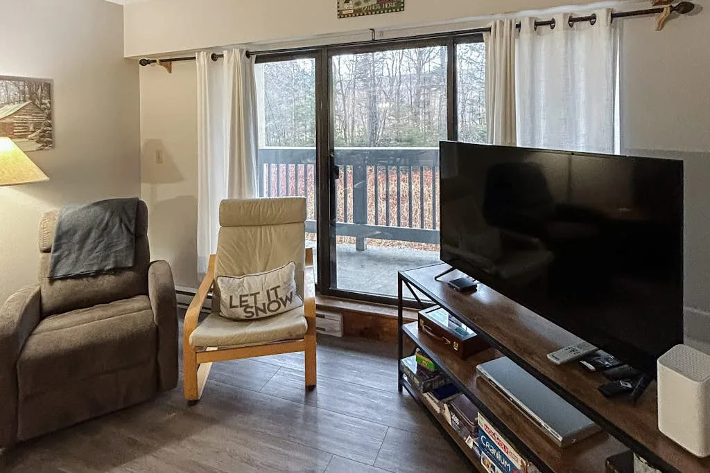 A living room with seating and a television beside a glass door overlooking a wooded view.