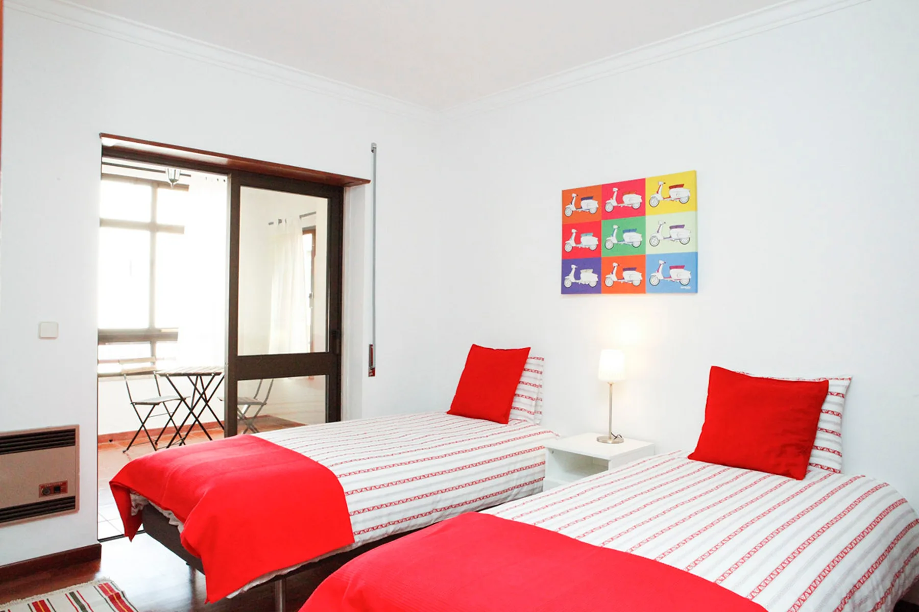 Un dormitorio luminoso con dos camas, ropa de cama roja y arte colorido en la pared.