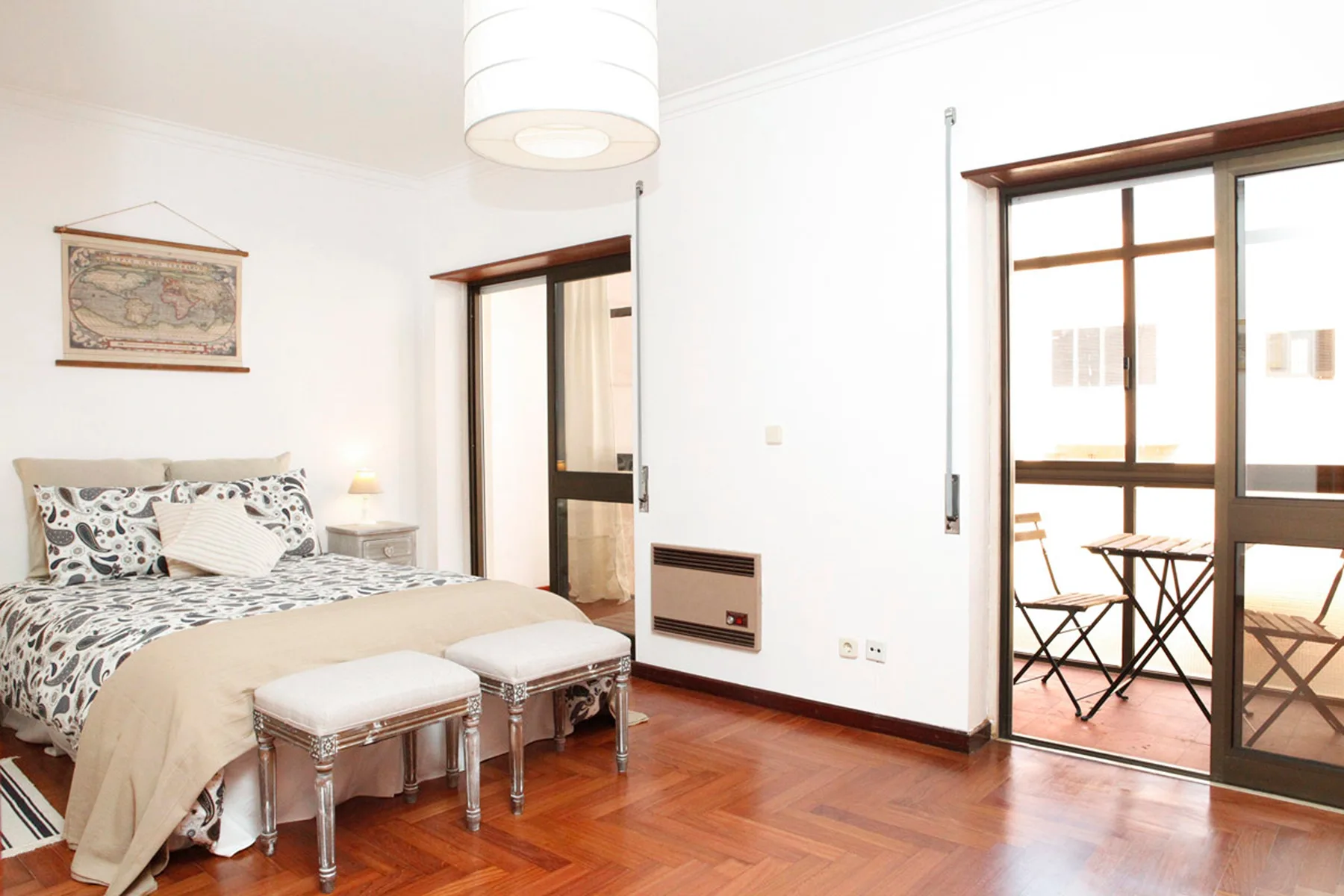 Un dormitorio luminoso con una cama, dos bancos tapizados y una puerta abierta a un balcón.