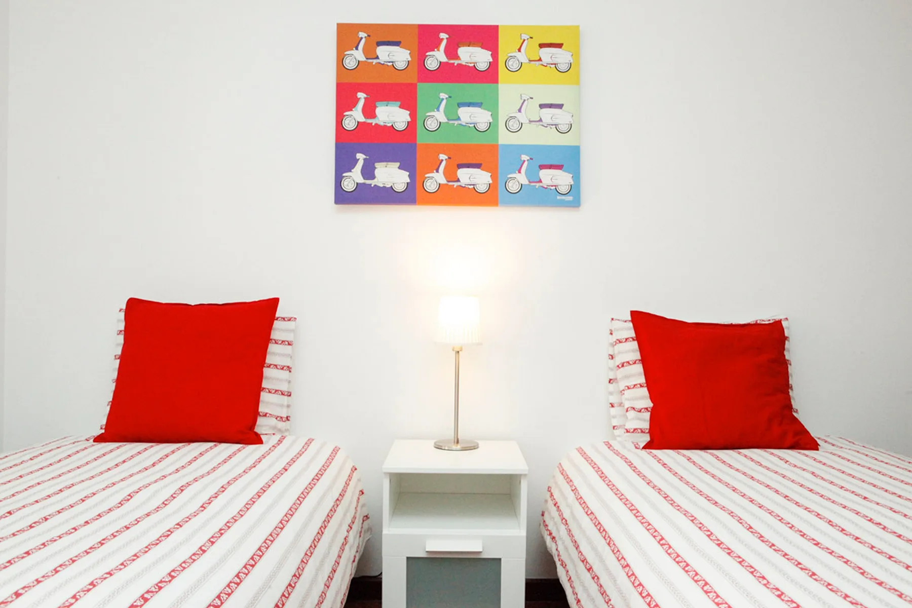 Un dormitorio con camas individuales, ropa de cama a rayas, cojines rojos y una lámpara.