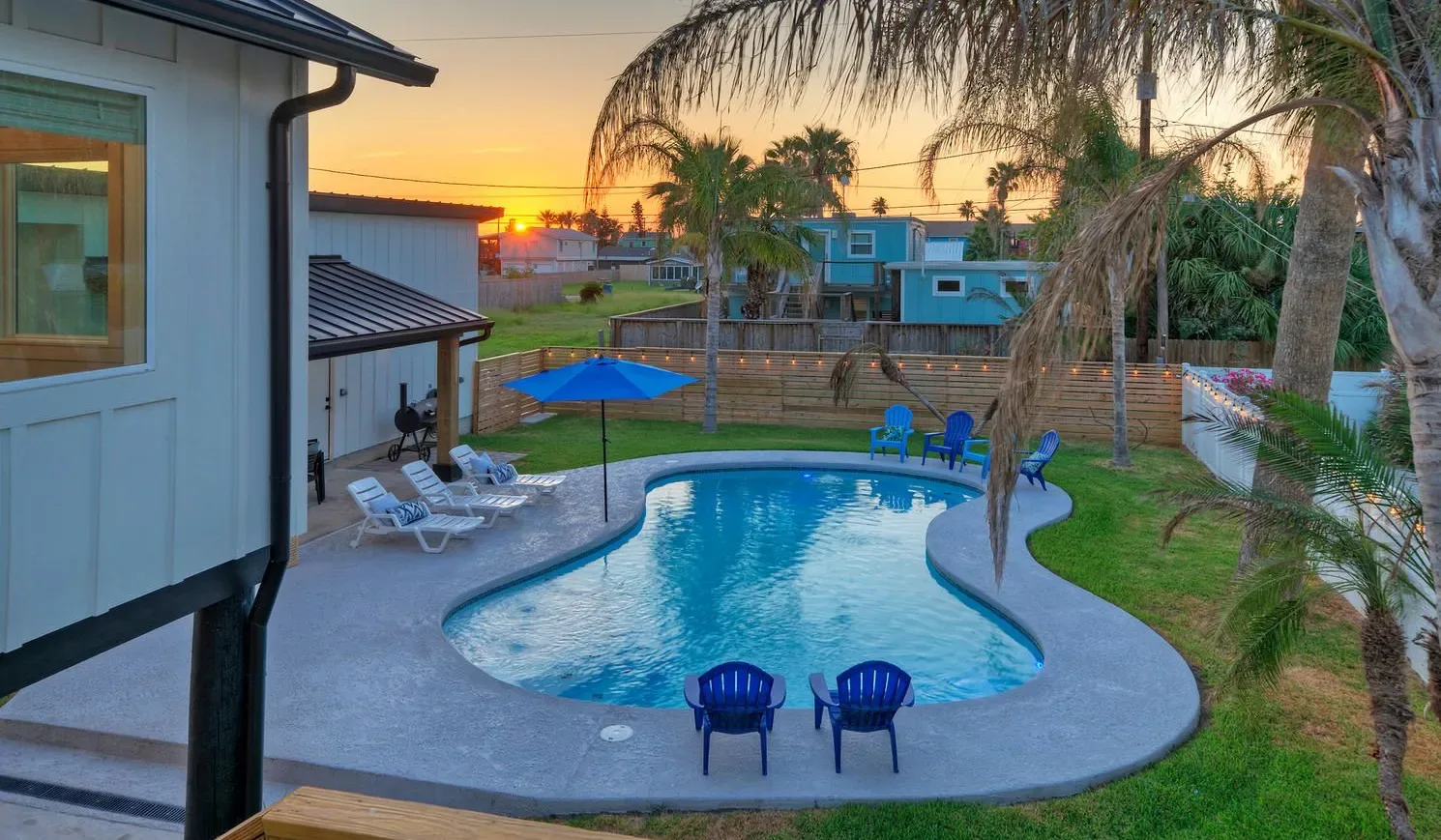 An outdoor swimming pool sits beside a patio with loungers and a blue umbrella.