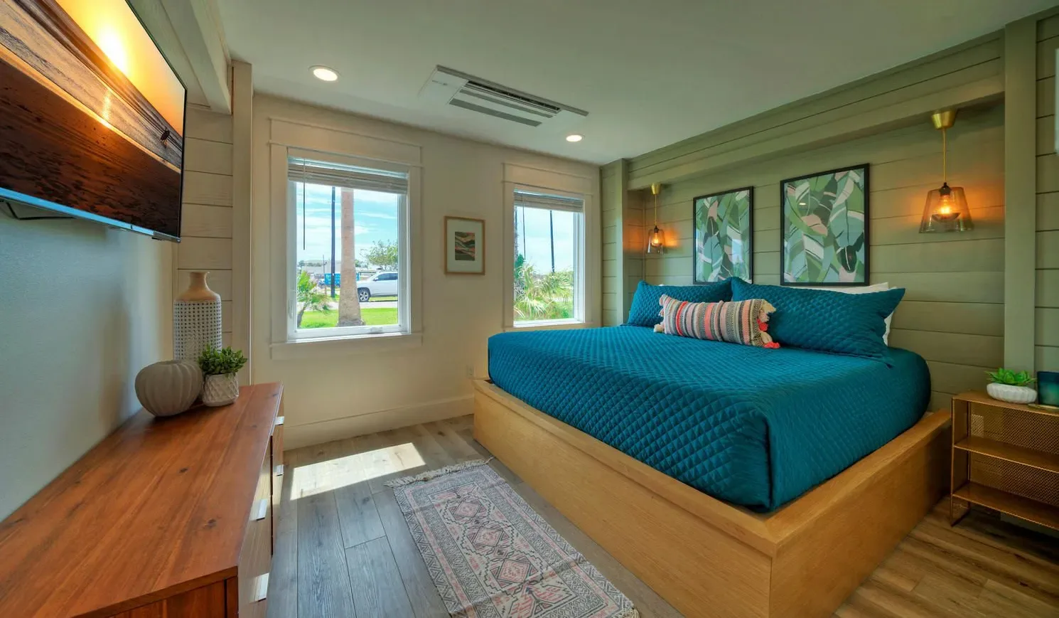 A bedroom with a teal bed, windows, and framed artwork on the walls.