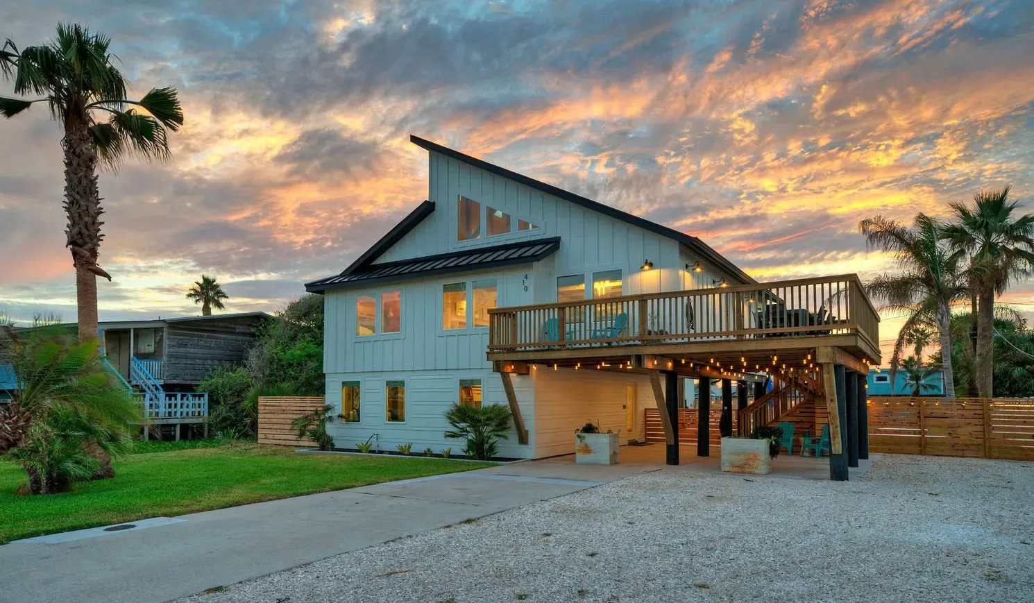 A coastal-style house with a raised wooden deck stands beneath a colorful sunset sky.
