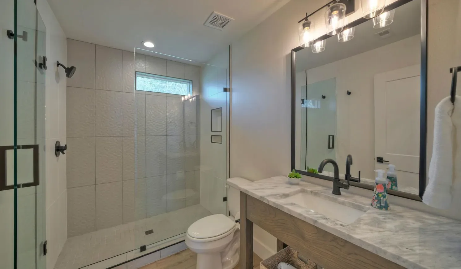 A bathroom with a glass shower, toilet, and long vanity beneath a large mirror.