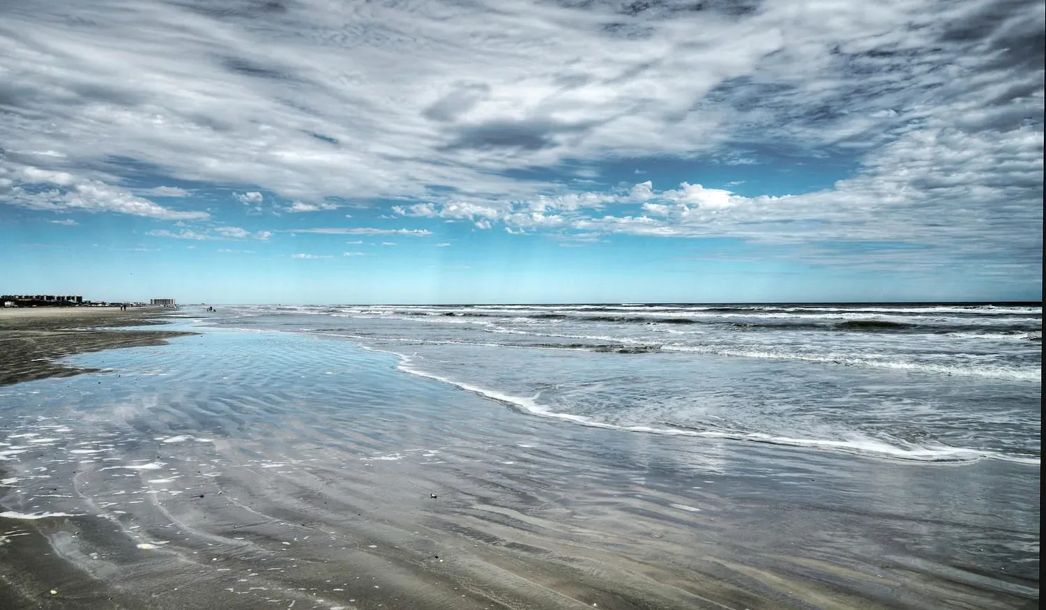 A broad beach shoreline meets shallow water beneath a cloudy sky.