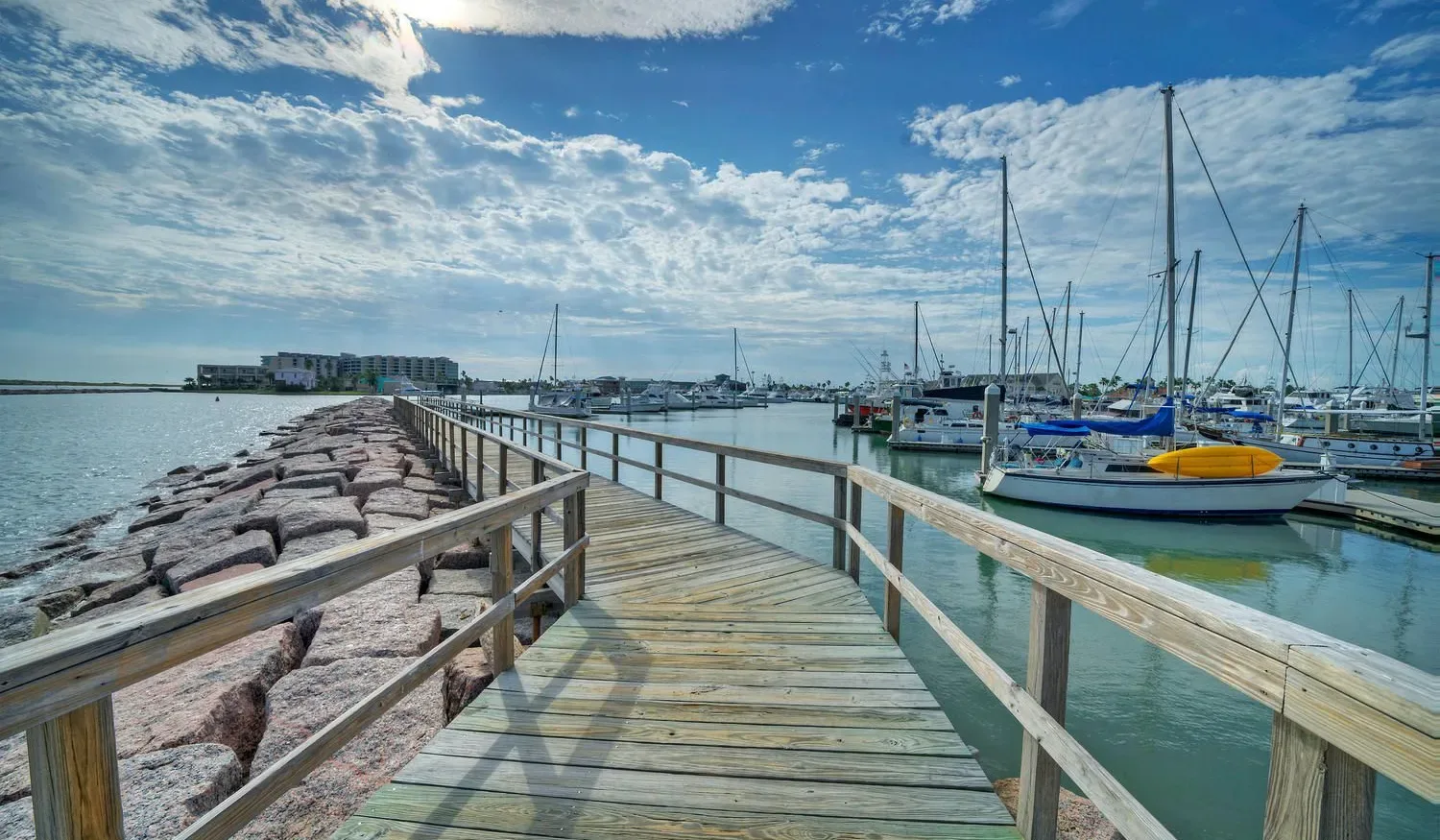A wooden walkway leads along a rocky shoreline beside a marina filled with sailboats.