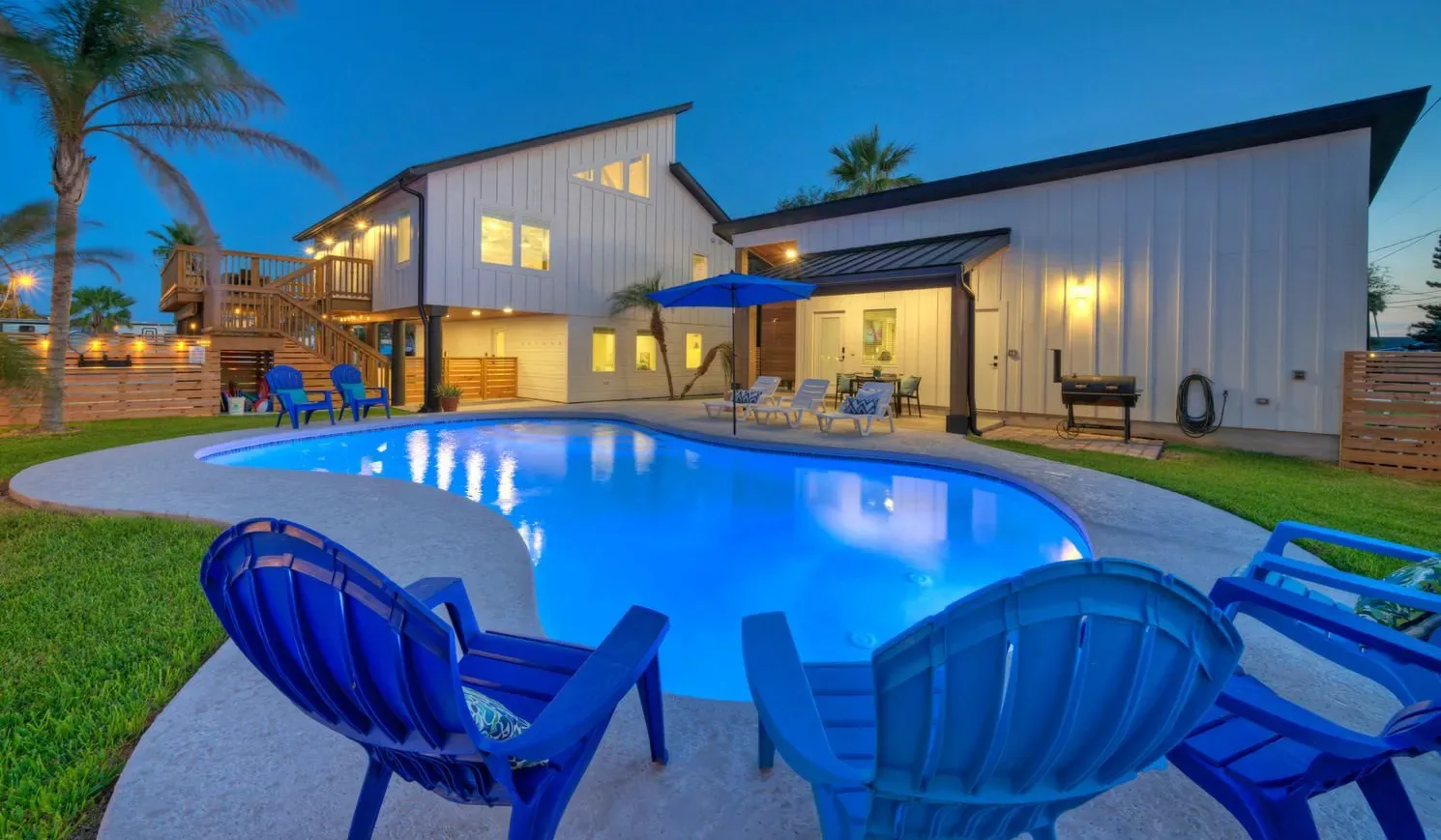 A lit modern house overlooks a swimming pool with blue outdoor seating.