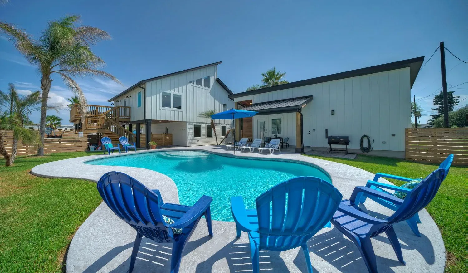 An outdoor swimming pool beside a modern house, with blue seating on the patio.