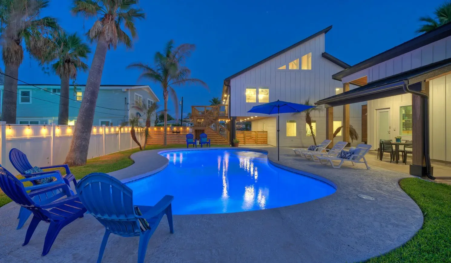 An illuminated swimming pool sits beside a modern house and outdoor seating at dusk.