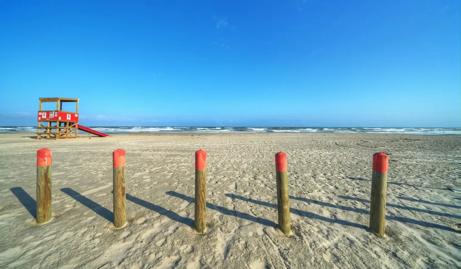 A sandy beach with a red lifeguard tower and wooden posts in the foreground.
