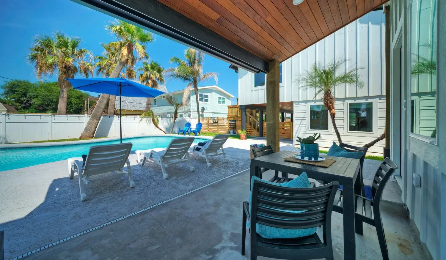 A covered patio with dining seating overlooks a swimming pool and palm trees.