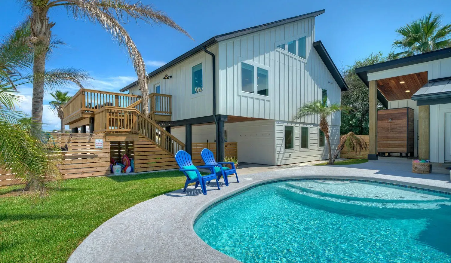 A white house stands beside a curved swimming pool, with blue chairs on the lawn.