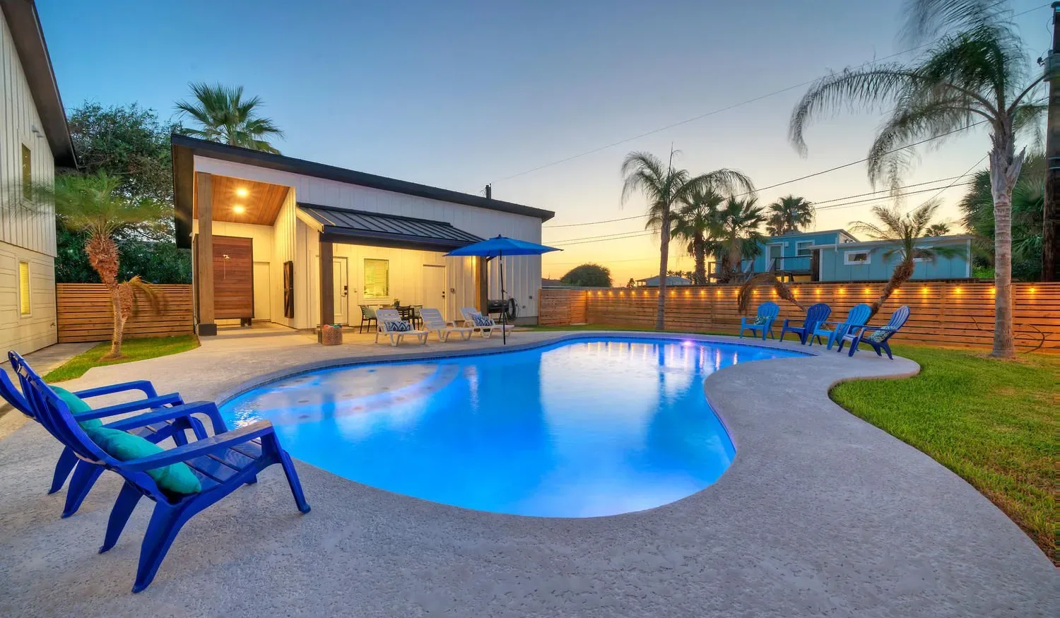 An outdoor swimming pool is surrounded by lounge seating and a lit patio.