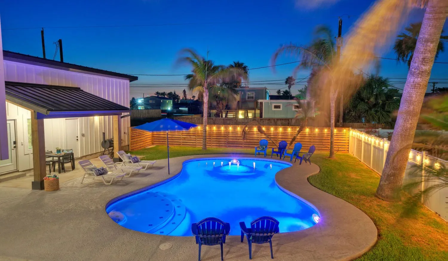 An illuminated backyard pool is surrounded by patio seating and palm trees at dusk.