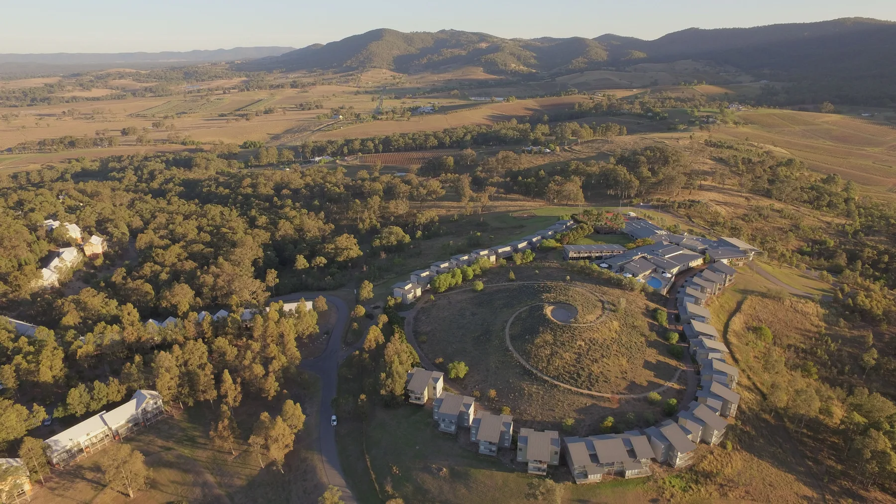 An aerial view shows a circular cluster of buildings amid trees and open fields.