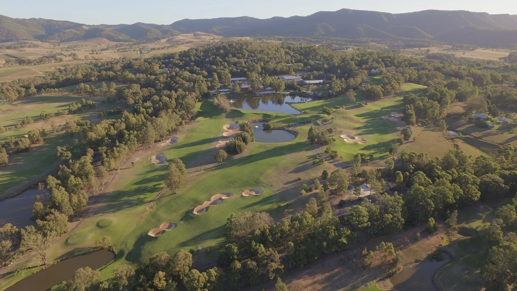 An aerial view of a golf course with ponds and wooded hills in the distance.