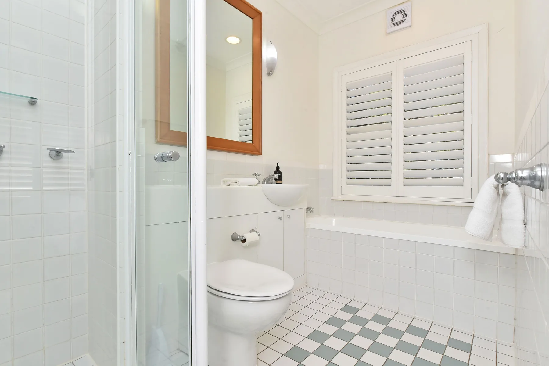 A bright bathroom with a shower, toilet, and window with shutters.