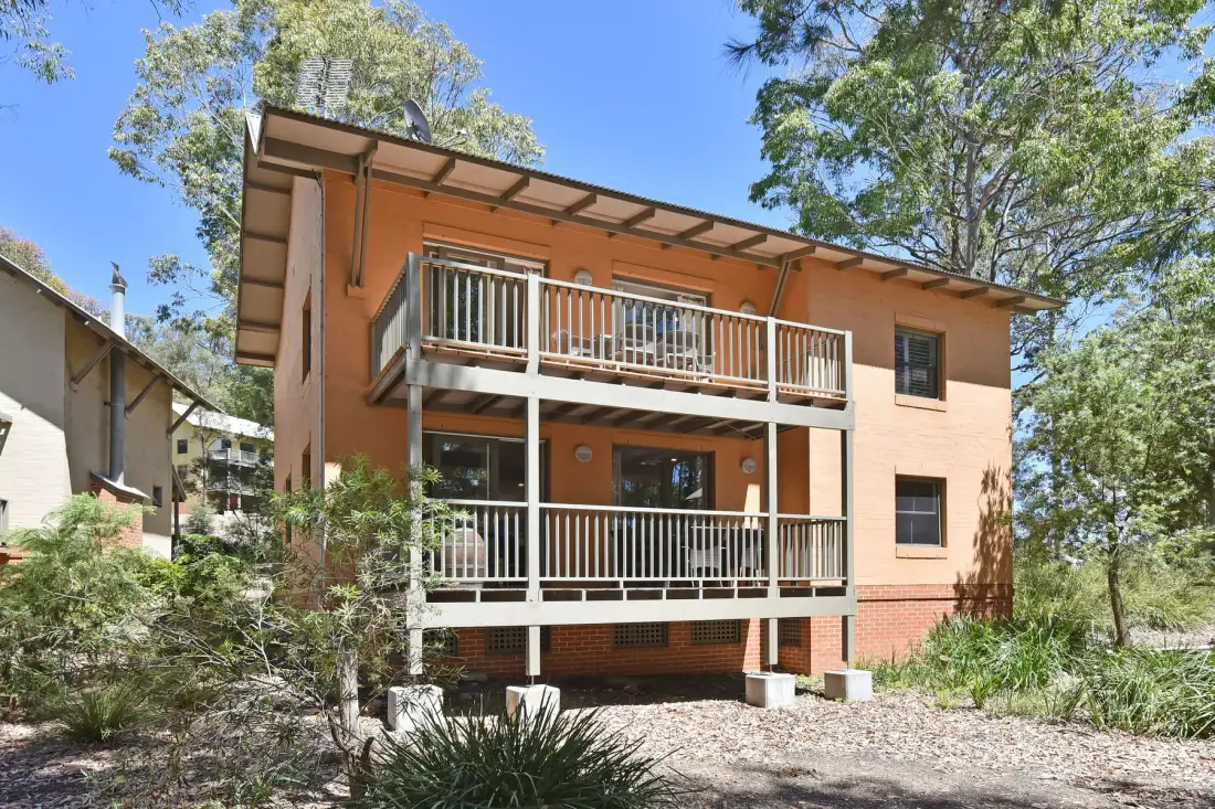 A two-story building with covered balconies stands among trees and shrubs.