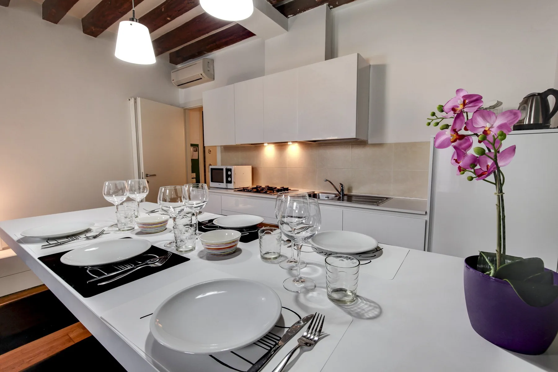 A dining table is set with dishes and glassware beside a kitchen.