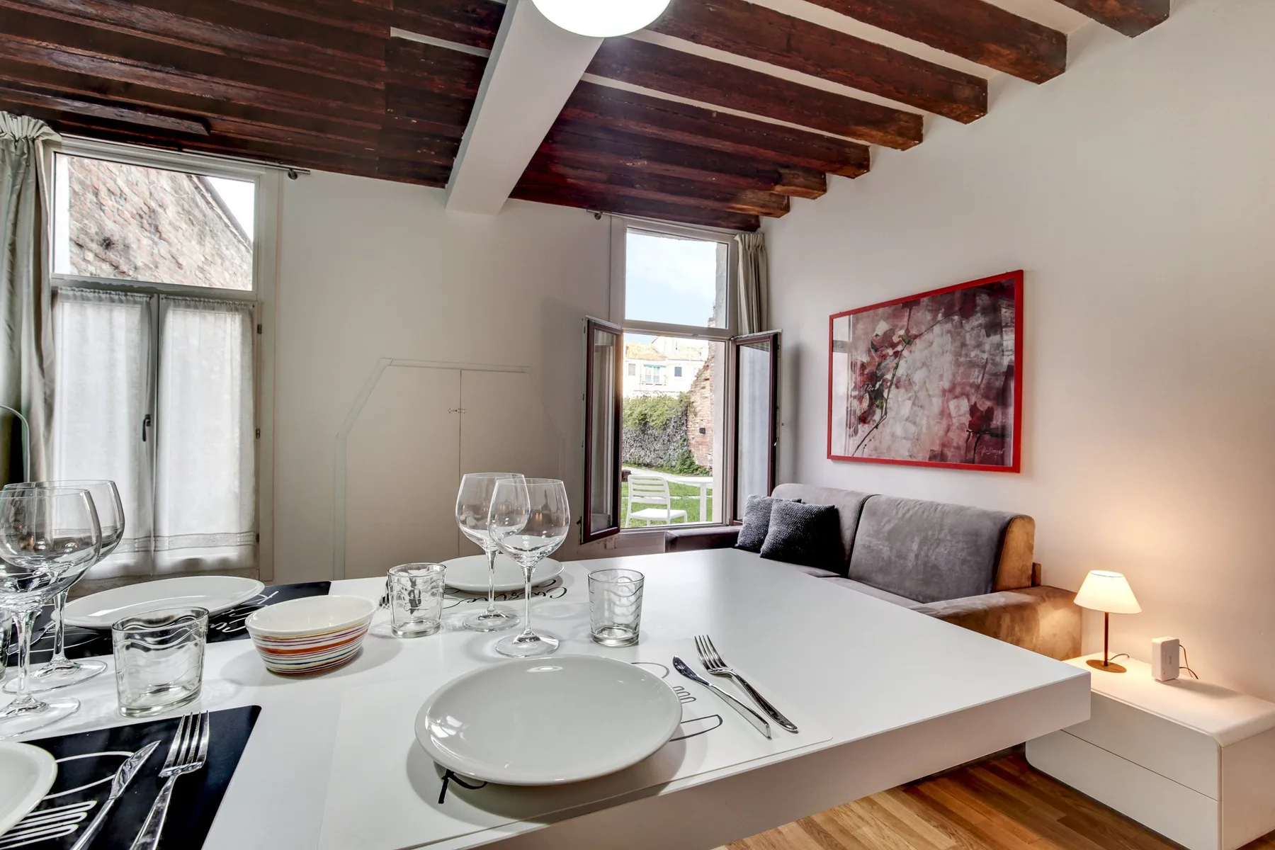 A dining area with a set table beside a gray sofa and large windows.
