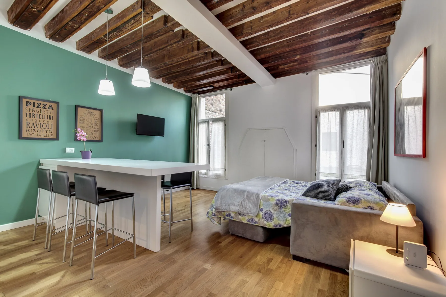 A bedroom with a bed, seating area, and counter beneath exposed wooden ceiling beams.