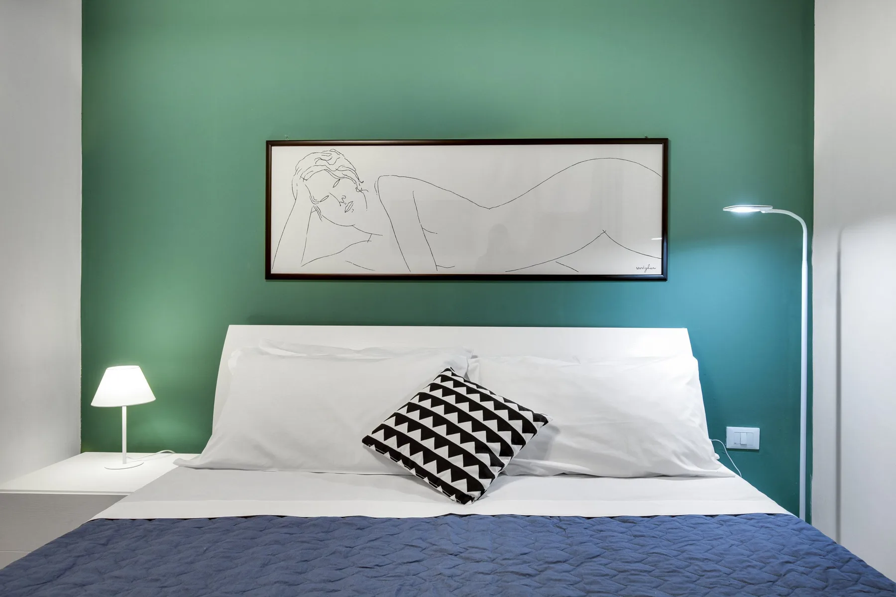A bedroom with a neatly made bed and framed artwork against a green wall.