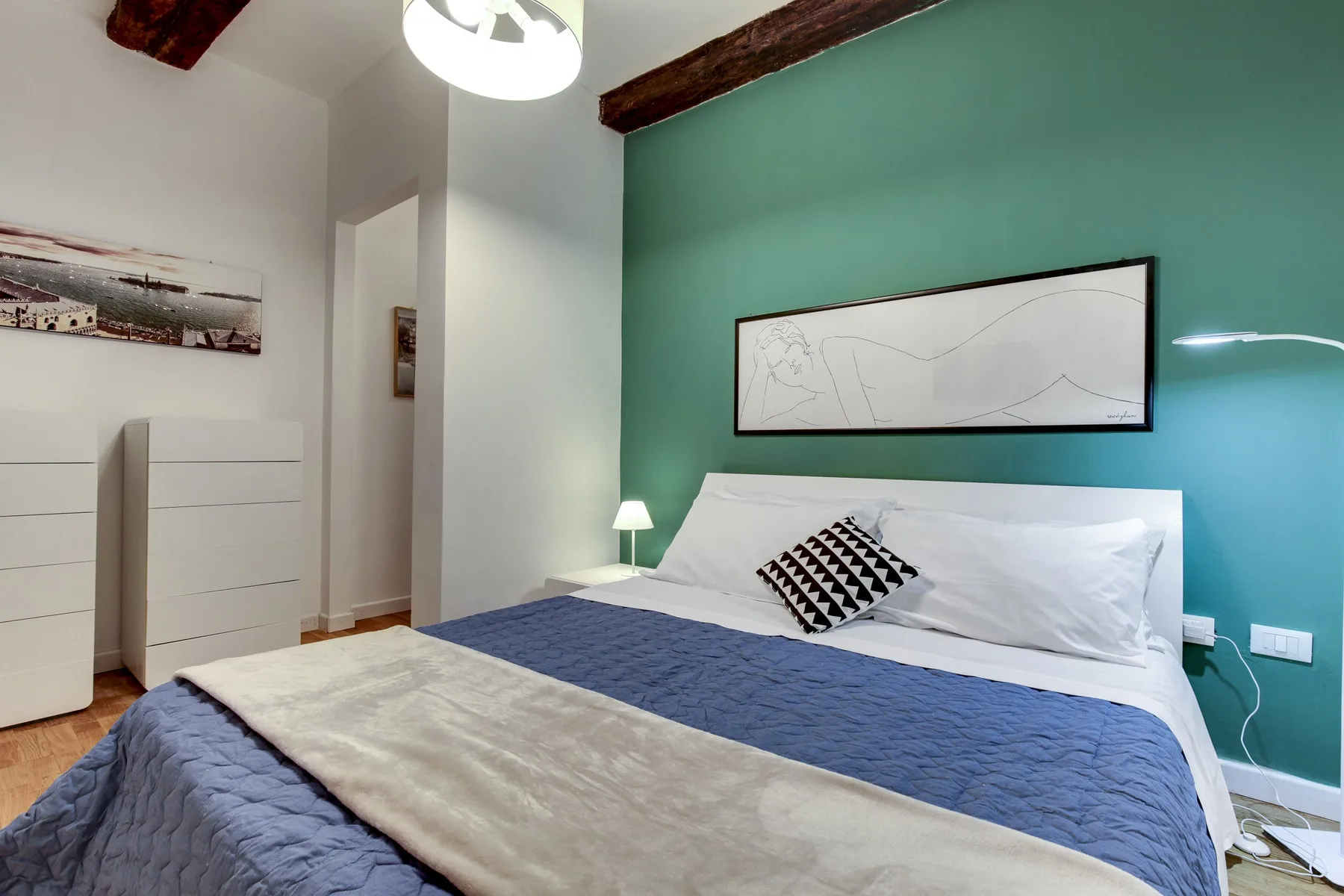 A bedroom with a bed against a teal wall and framed artwork above it.
