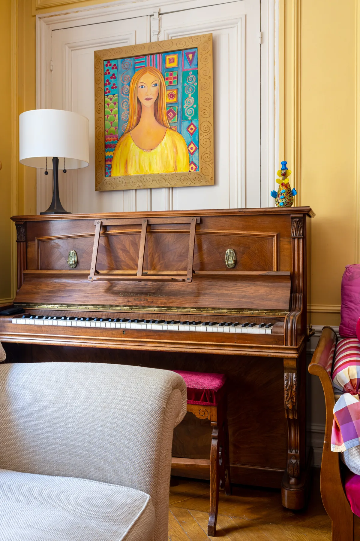 A wooden piano stands against a yellow wall beneath a colorful portrait.