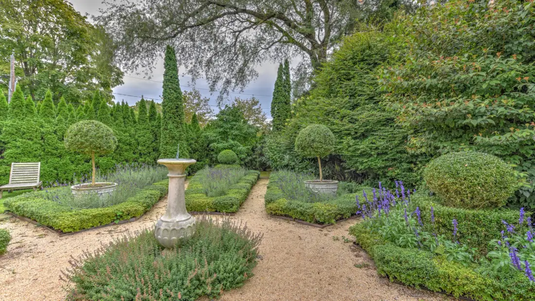 Un jardín ajardinado con senderos de grava, setos recortados y una fuente central de piedra.