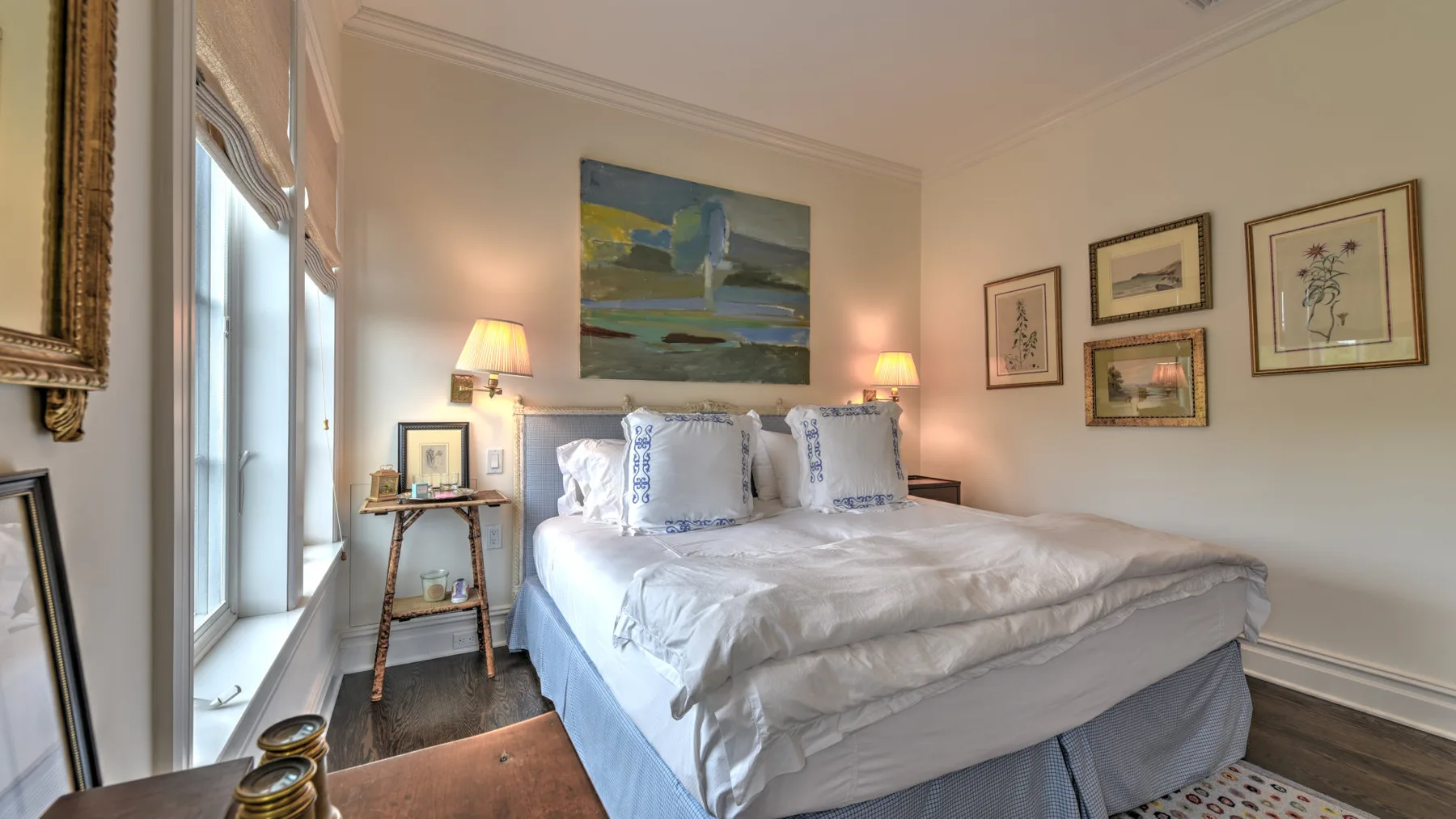 Un dormitorio con una cama bien tendida junto a una ventana alta y cuadros enmarcados.