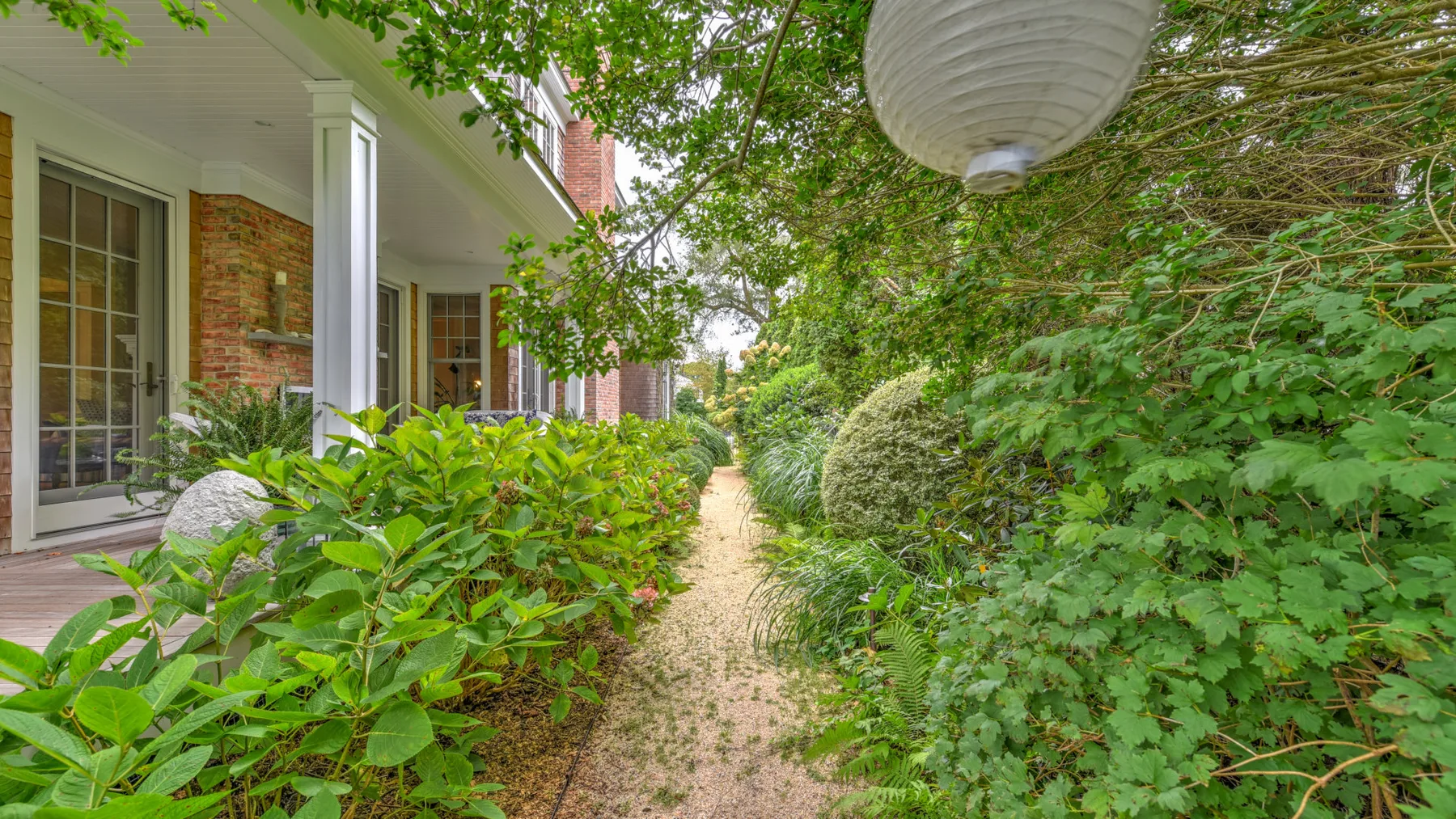 Un sendero de jardín pasa entre plantas frondosas junto a una casa de ladrillo.