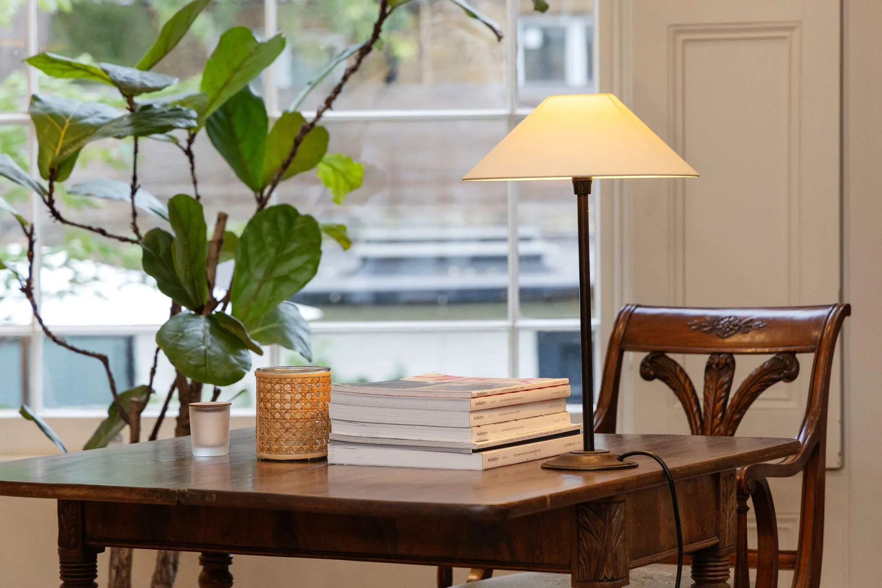 A wooden table with books and a lit lamp beside a window.