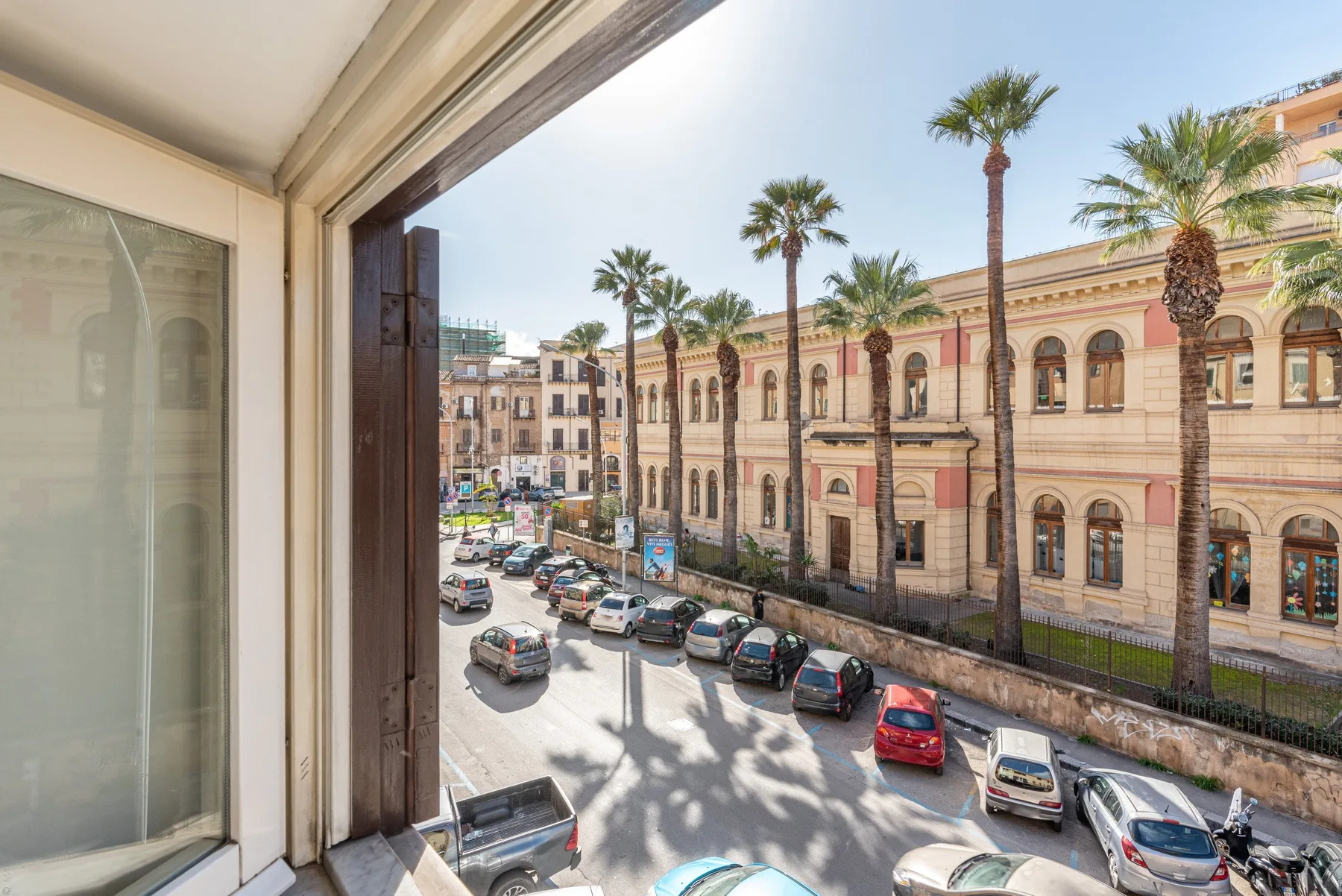Vista de una calle desde una ventana, con coches estacionados y palmeras.