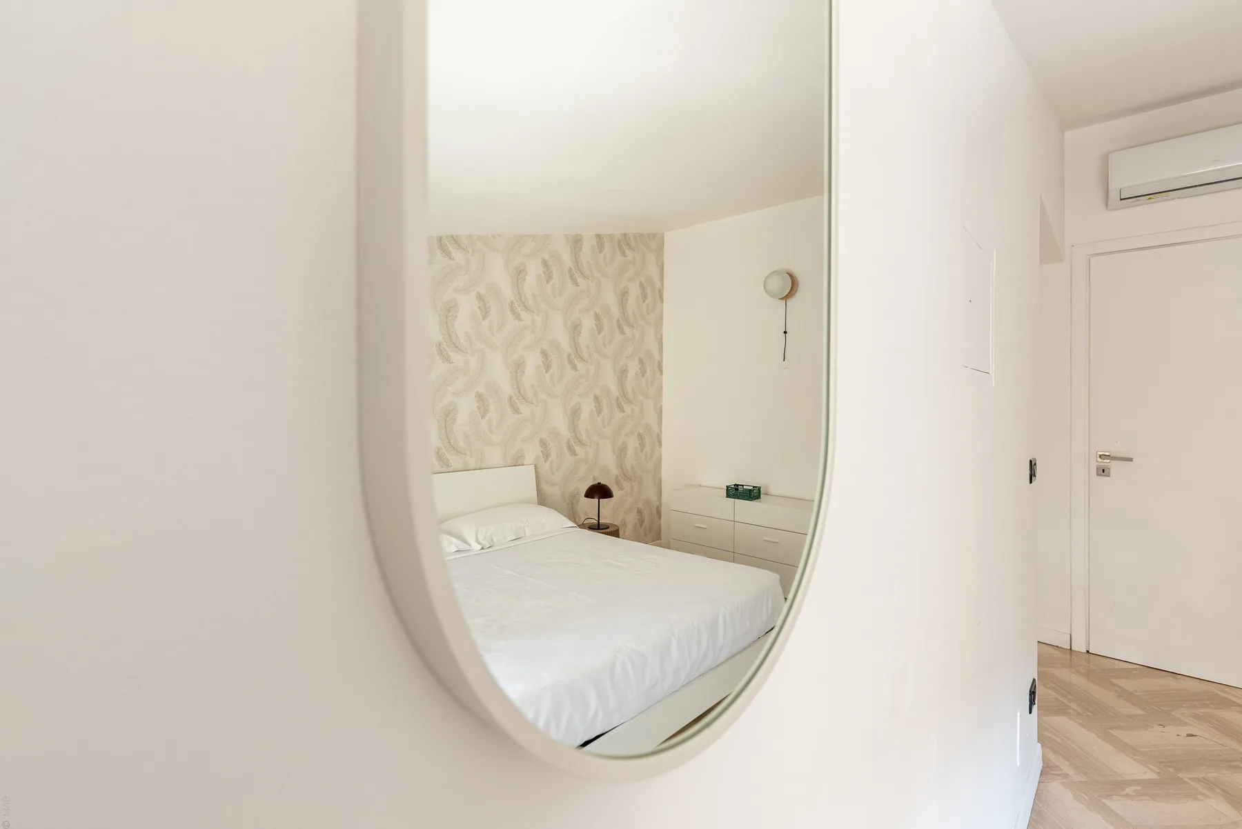 Dormitorio reflejado en un gran espejo de pared, con papel pintado estampado detrás de la cama.