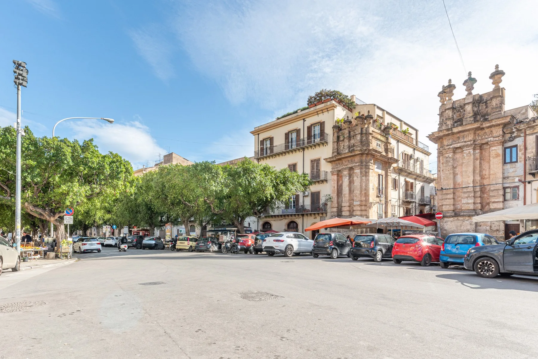 Una plaza soleada con coches estacionados, árboles frondosos y edificios históricos de piedra.