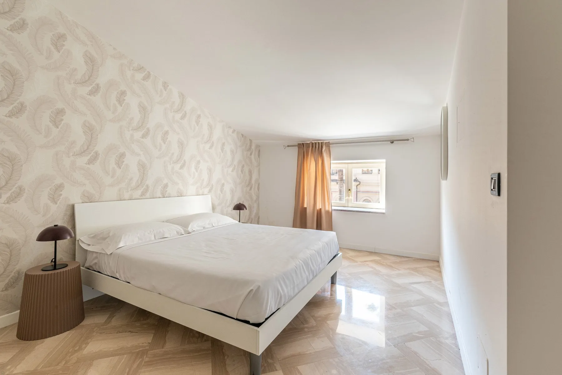 Un dormitorio luminoso con una cama grande, papel tapiz estampado y una ventana con cortinas.