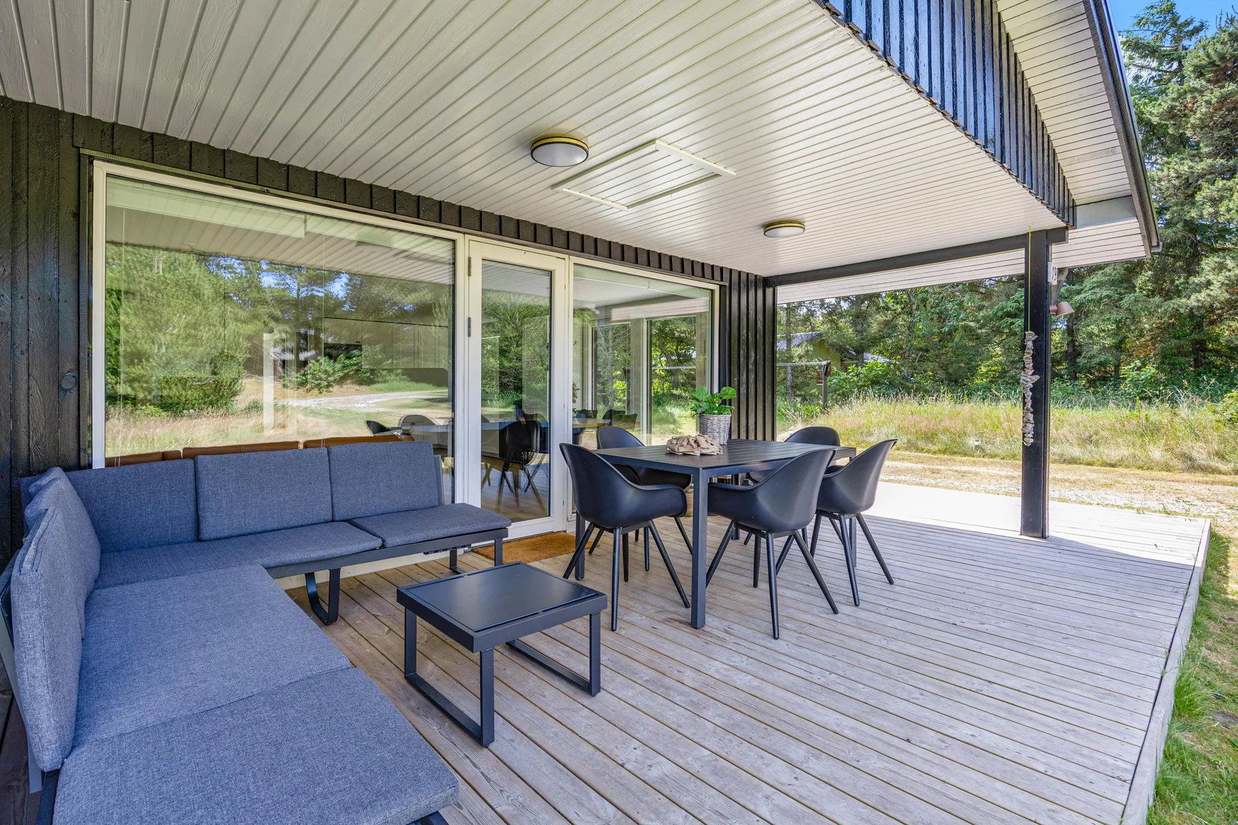 Una zona exterior cubierta para sentarse, con una mesa y sillas junto a grandes ventanas.