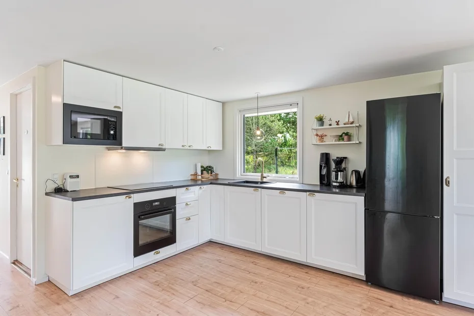Una cocina luminosa con armarios blancos, encimeras oscuras y una ventana con vista a la vegetación.