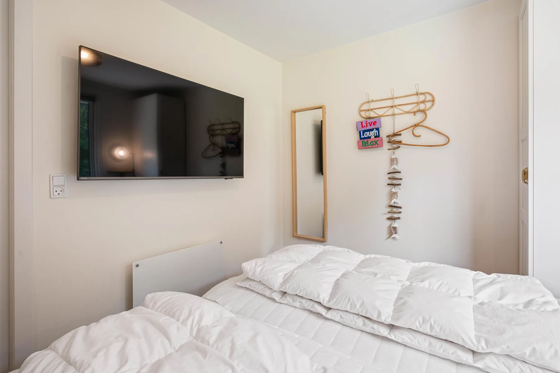 Un dormitorio con ropa de cama blanca, un televisor montado en la pared y un espejo estrecho.