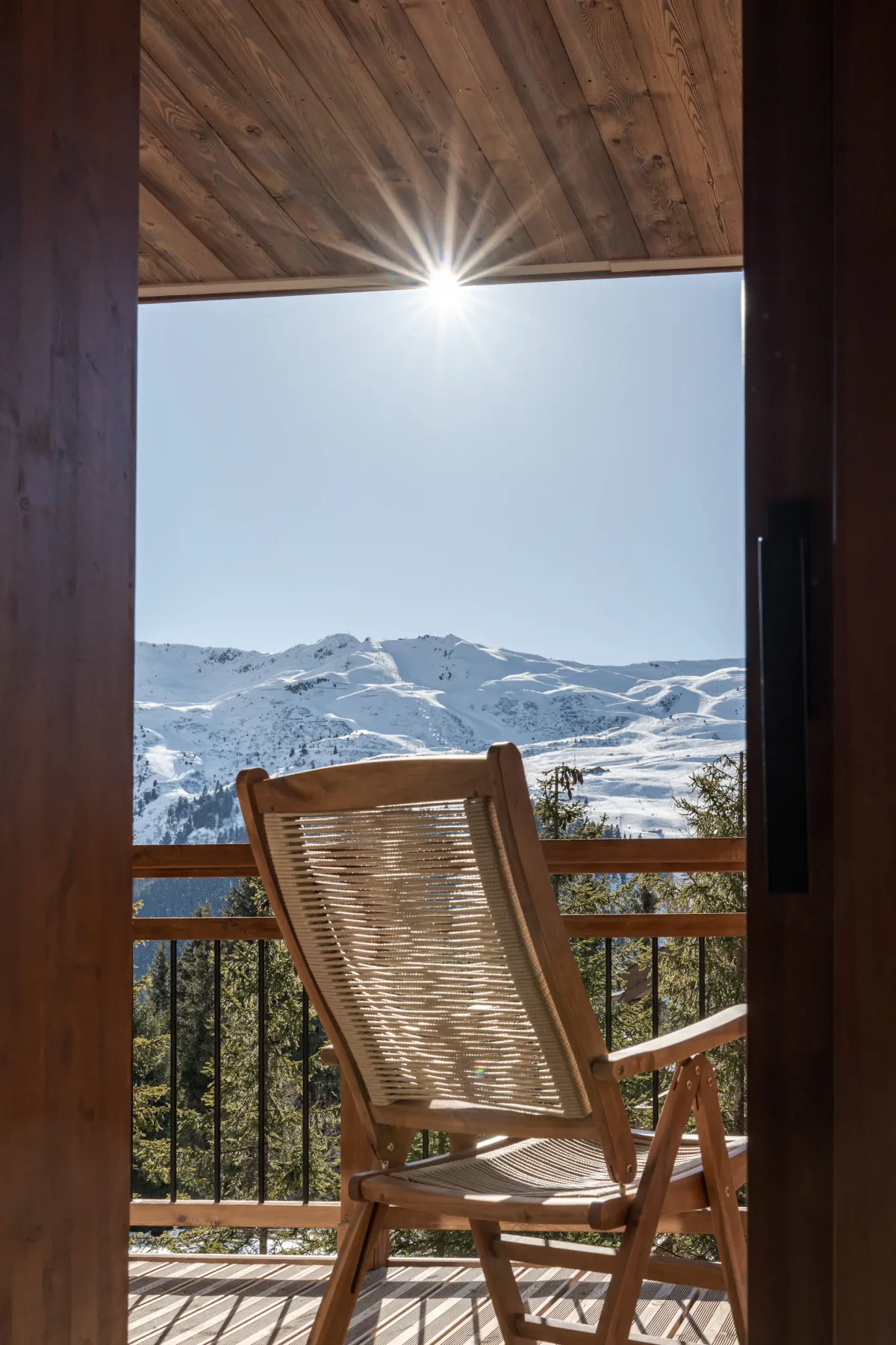 Un balcón de madera con una silla reclinable ofrece vistas a montañas nevadas bajo un cielo luminoso.