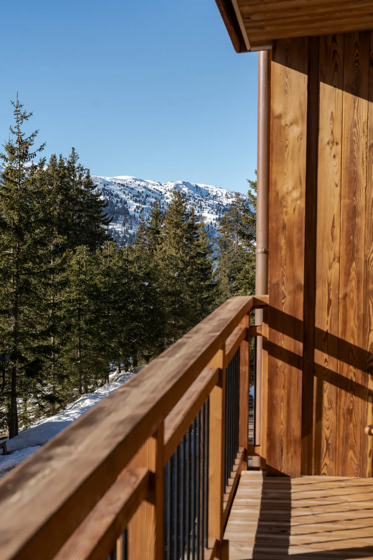 Un balcón de madera ofrece vistas a árboles de hoja perenne y montañas nevadas bajo un cielo azul despejado.