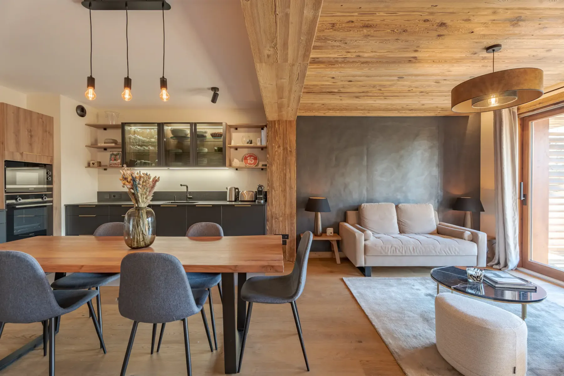 Cocina moderna con armarios de madera, una encimera negra y una mesa de comedor con seis sillas grises. Sobre la mesa cuelgan lámparas. En los estantes se exhiben piezas de cristalería y un plato rojo.