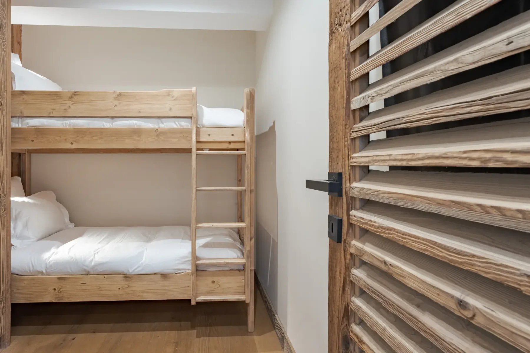 Literas de madera con ropa de cama blanca situadas contra una pared. En primer plano, una puerta de madera con lamas horizontales está parcialmente abierta.