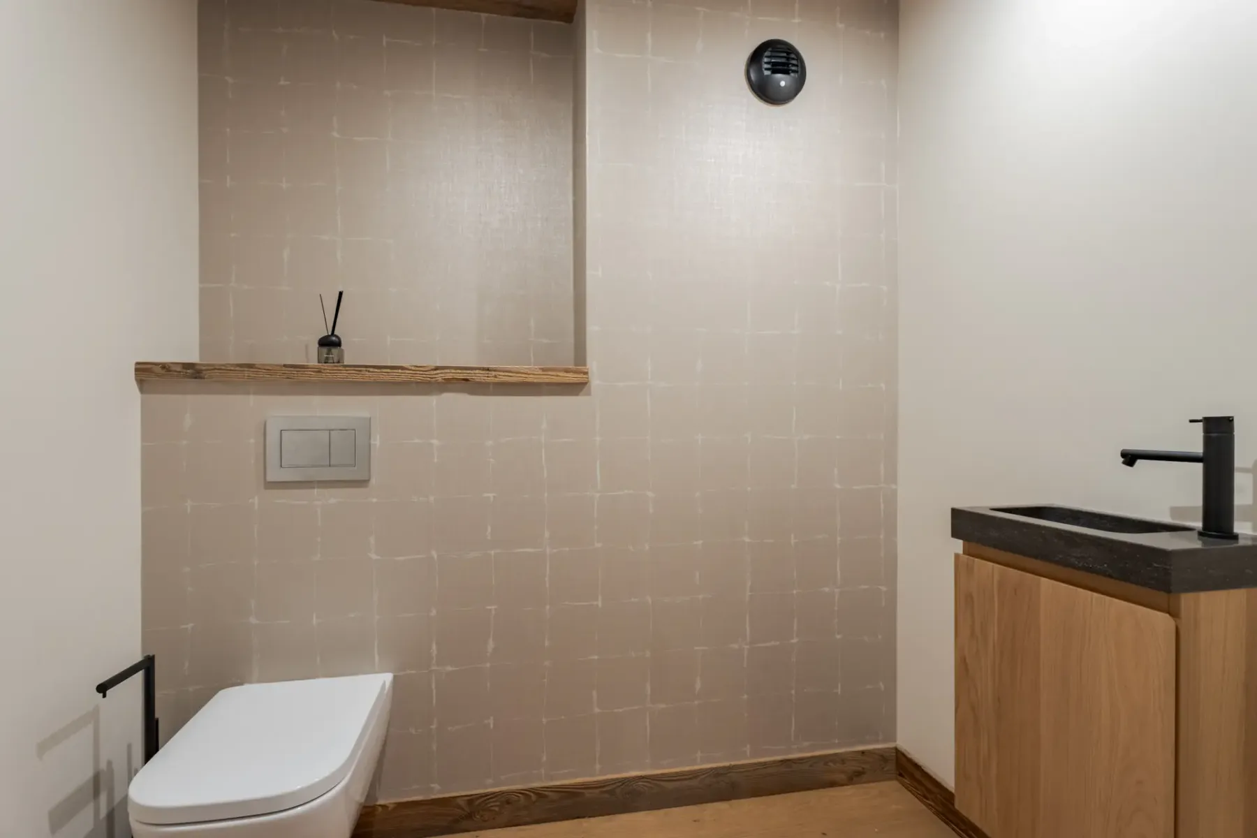Baño minimalista con un inodoro suspendido, un pequeño grifo negro para el lavabo y muebles de madera. Sobre el inodoro hay un estante con un difusor y una rejilla de ventilación. Las paredes tienen un sutil patrón de cuadrícula.