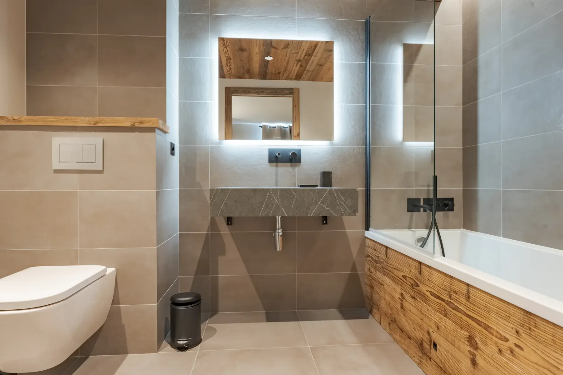 Baño moderno con azulejos beige, un inodoro suspendido, un espejo retroiluminado sobre un lavabo y una bañera con un panel de madera.