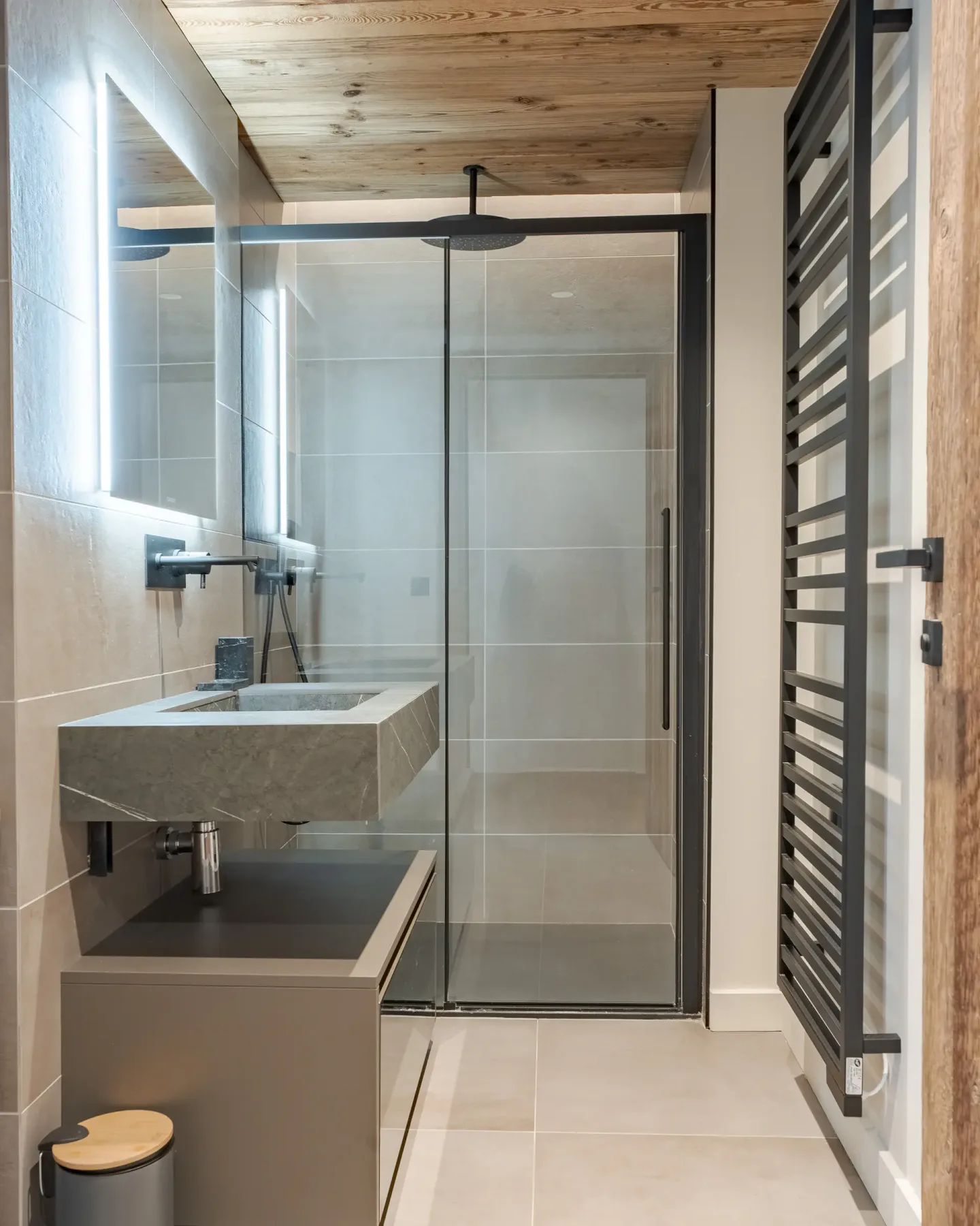 Baño moderno con una ducha con mampara de cristal, un espejo iluminado sobre un lavabo de diseño elegante, techo de madera y un cesto para la ropa. Decoración minimalista en tonos neutros y suelo de baldosas.