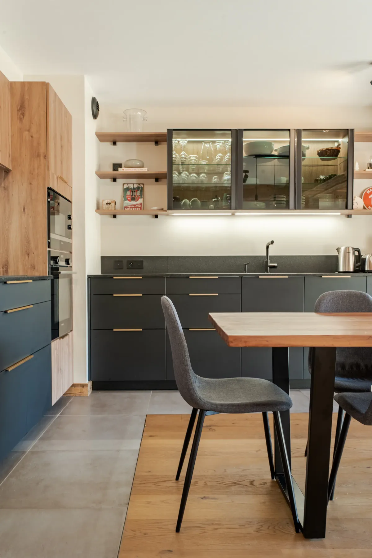 Cocina moderna con armarios de madera, una encimera negra y una mesa de comedor con seis sillas grises. Sobre la mesa cuelgan lámparas. En los estantes se exhiben piezas de cristalería y un plato rojo.