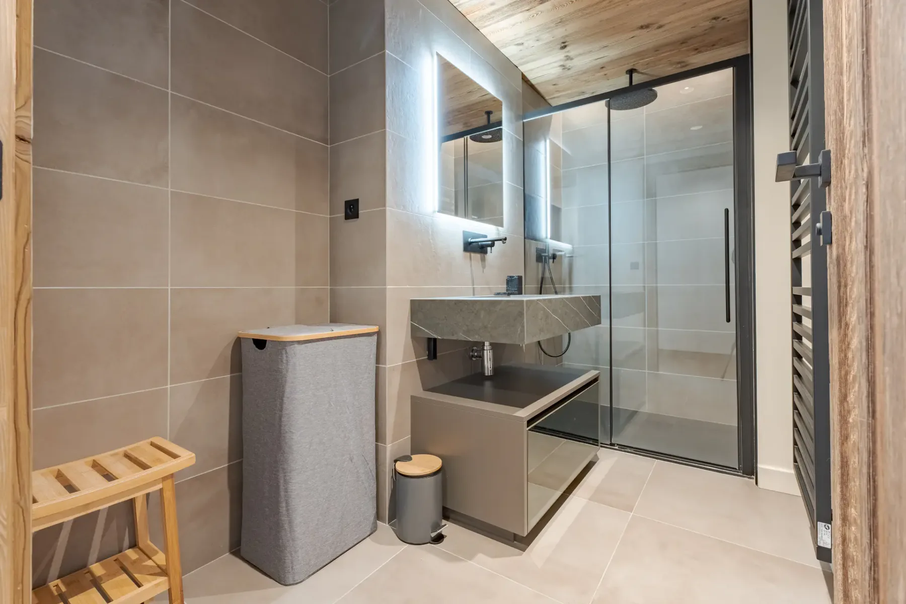 Baño moderno con una ducha con mampara de cristal, un espejo iluminado sobre un lavabo de diseño elegante, techo de madera y un cesto para la ropa. Decoración minimalista en tonos neutros y suelo de baldosas.