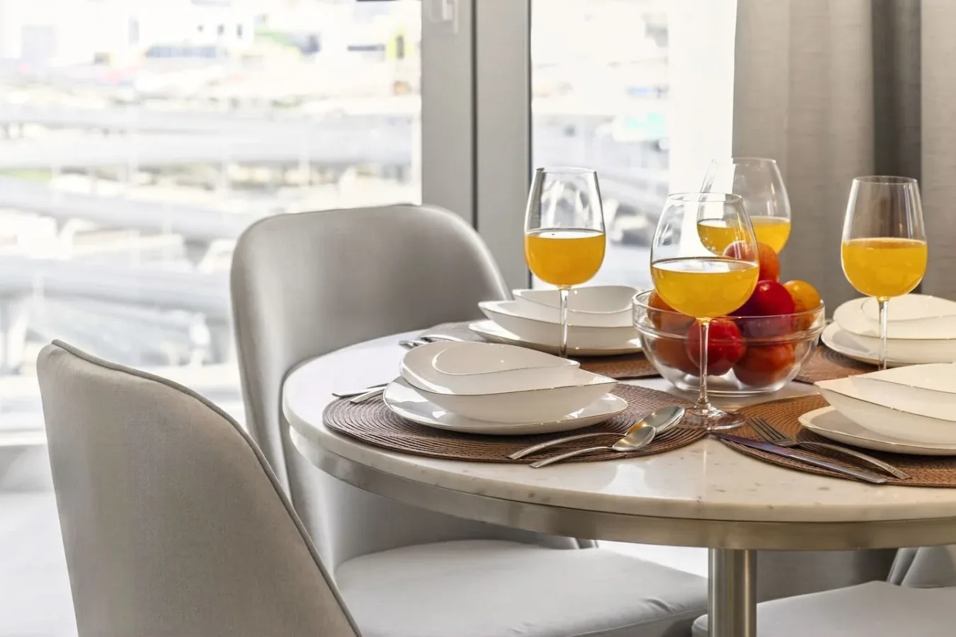 Una mesa de comedor puesta con platos, vasos de jugo y un frutero junto a una ventana.